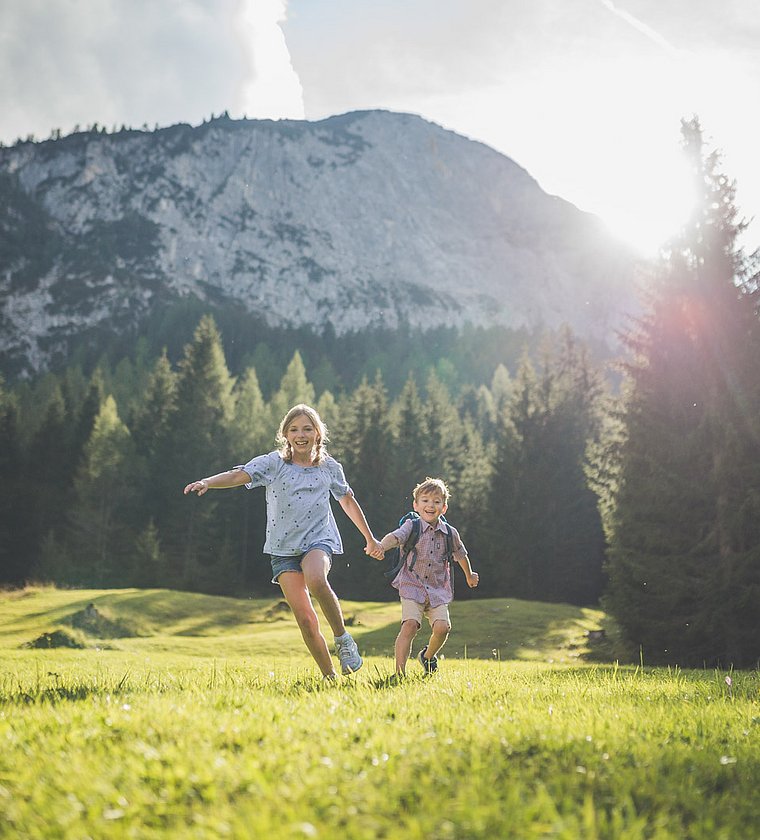 Two children run across a sunny alpine meadow with mountains in the background