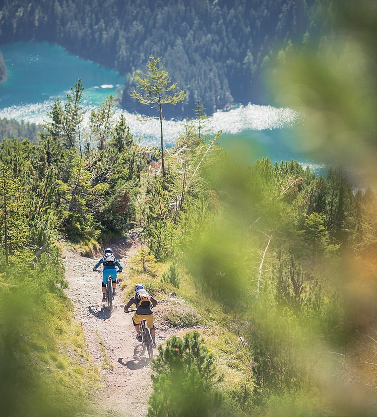 Two mountain bikers on a forest trail overlooking the turquoise Heiterwang Lake
