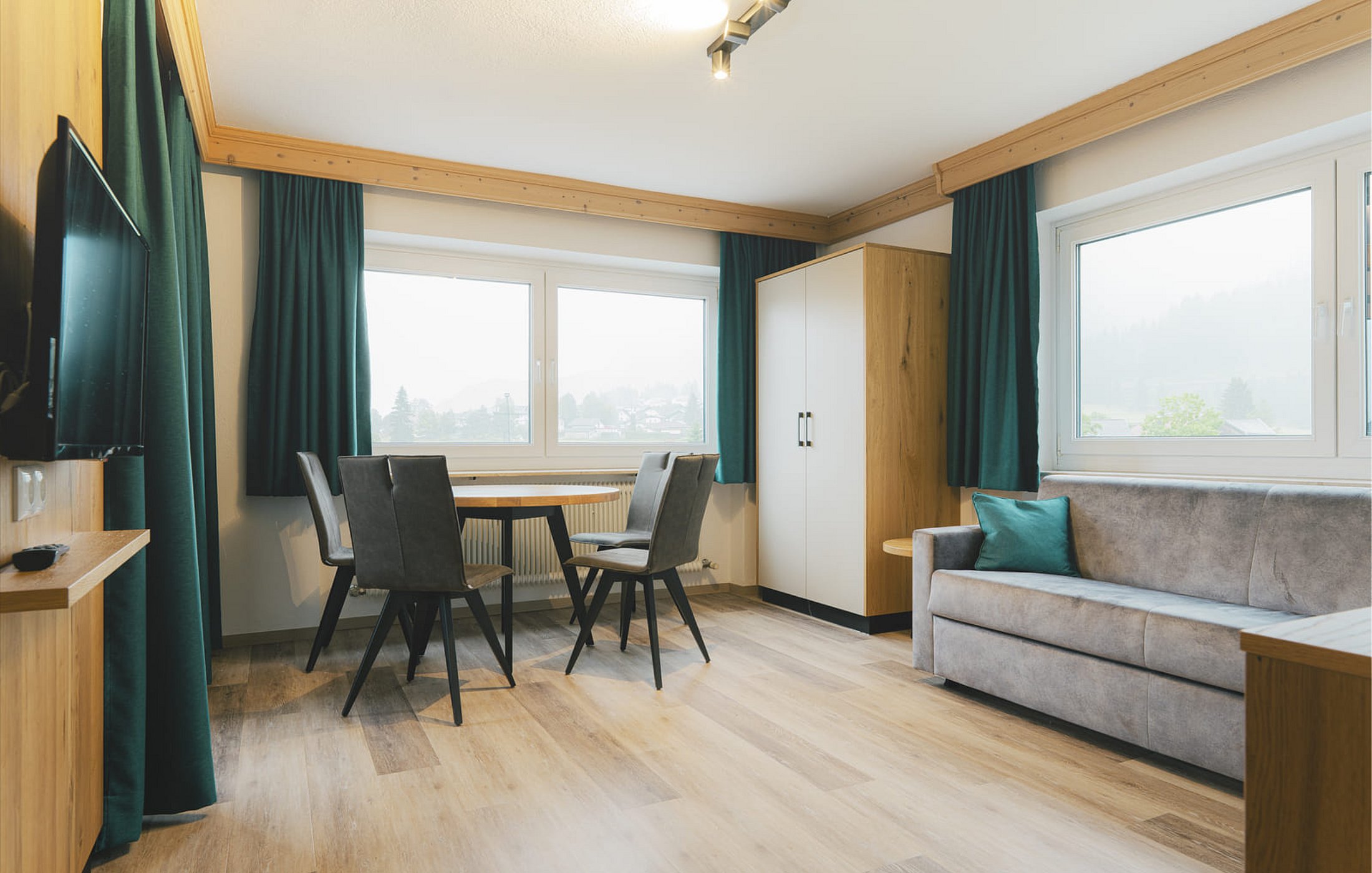 Wohnzimmer in der Ferienwohnung mit Holztisch, Sofa, Kleiderschrank und grünen Vorhängen